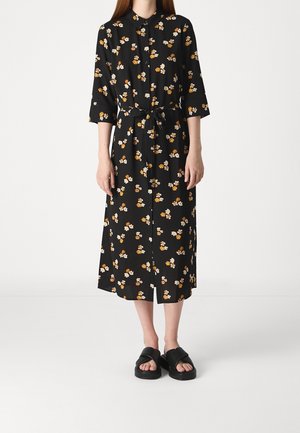 Frau trägt ein schwarzes Midikleid mit weißem und orangefarbenem Blumenmuster, Dreiviertelärmeln, Bindeband in der Taille und schwarzen Sandalen mit offenen Zehen.