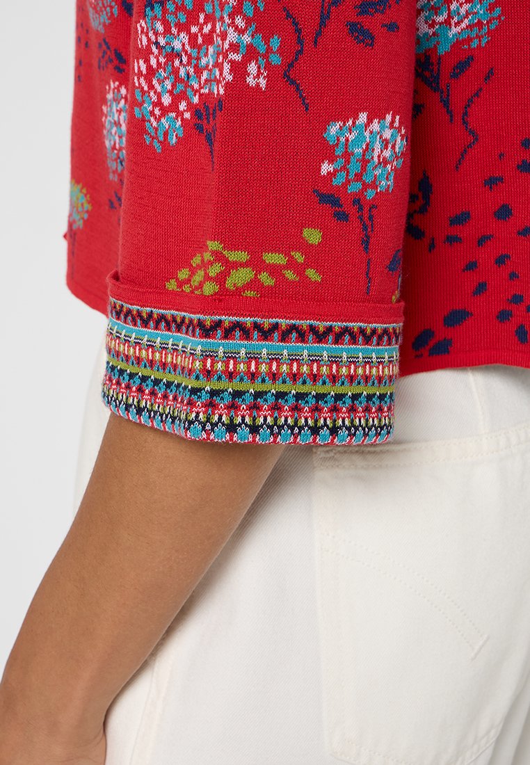 Czerwona dzianinowa rękawica z kwiatowymi wzorami i wielokolorowym, misternie zdobionym mankietem, noszona z białymi spodniami, z widoczną częścią tylnej kieszeni.