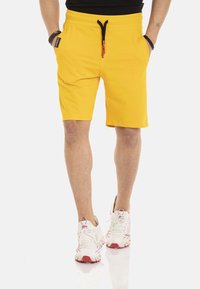Gele shorts van zachte stof, voorzien van een elastische tailleband met een zwarte trekkoord en zijzakken. Draagt witte sneakers.
