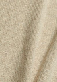 Närbild av beige stickad tyg som visar ett mjukt, fint strukturerat ullmönster med mjuka veck och en slät yta.