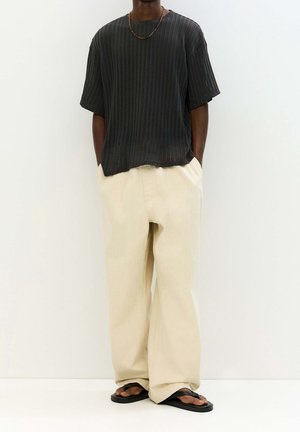 Man wearing a black ribbed short-sleeve shirt, loose beige pants, black flip-flops, and a gold chain necklace against a white background.