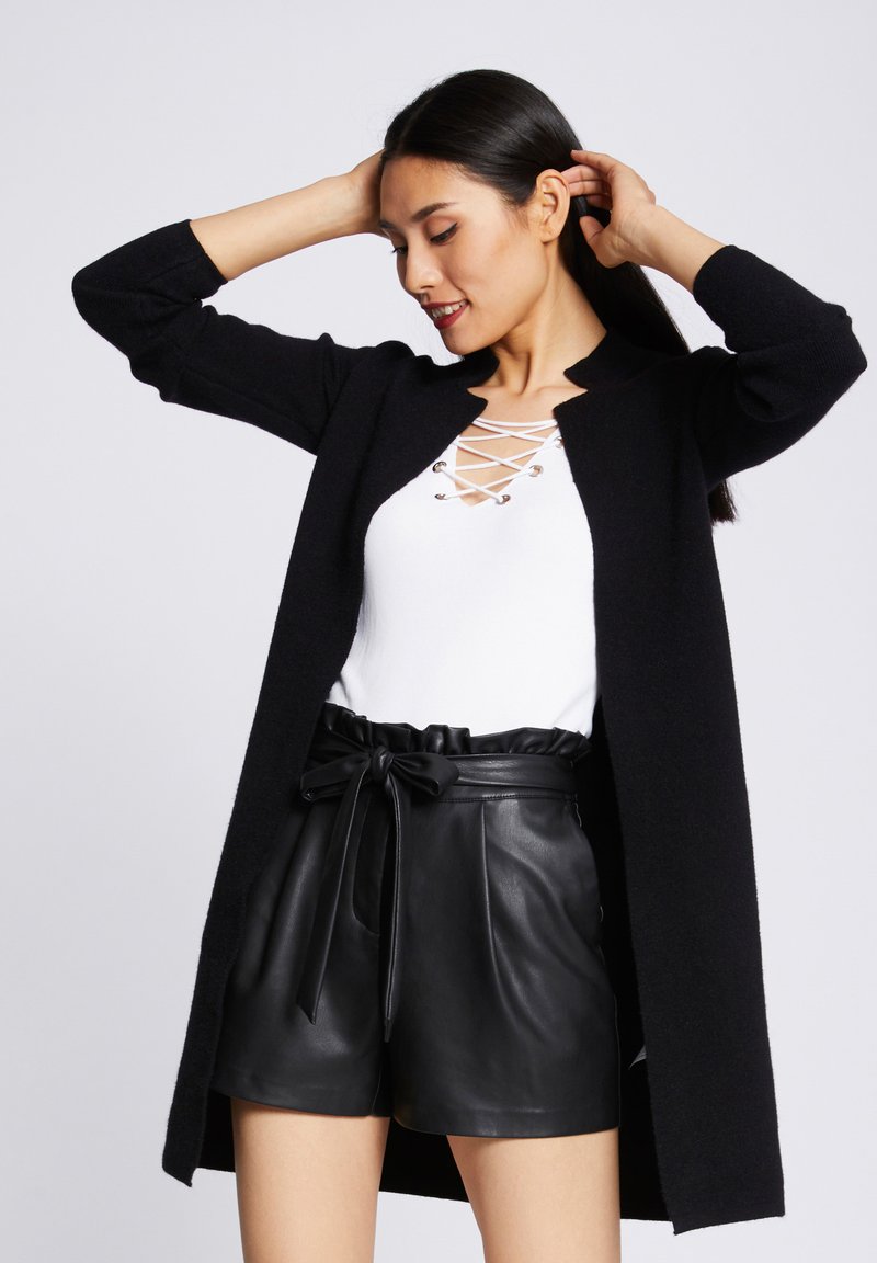 Long cardigan noir en texture côtelée porté sur un haut blanc avec lacets et œillets argentés, associé à un short en cuir noir taille haute.