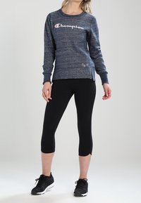 Donna che indossa una felpa navy Champion, leggings neri a tre quarti e sneakers nere, in piedi contro uno sfondo neutro.