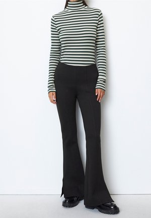 Kobieta ubrana w czarne dzwony, czarne masywne buty oraz zielono-biały, paskowany, dopasowany golf z długim rękawem, stoi na białym tle.