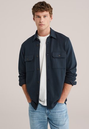 Ung mand med krøllet hår iført en mørkeblå skjorte med knapper over en hvid T-shirt og lyseblå jeans, hænderne i lommerne, neutral baggrund.