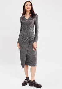 Abito argento dalla texture con dettaglio frontale attorcigliato e maniche lunghe, caratterizzato da una gonna avvolgente drappeggiata e un orlo all'altezza del ginocchio. Scarpe nere chunky.