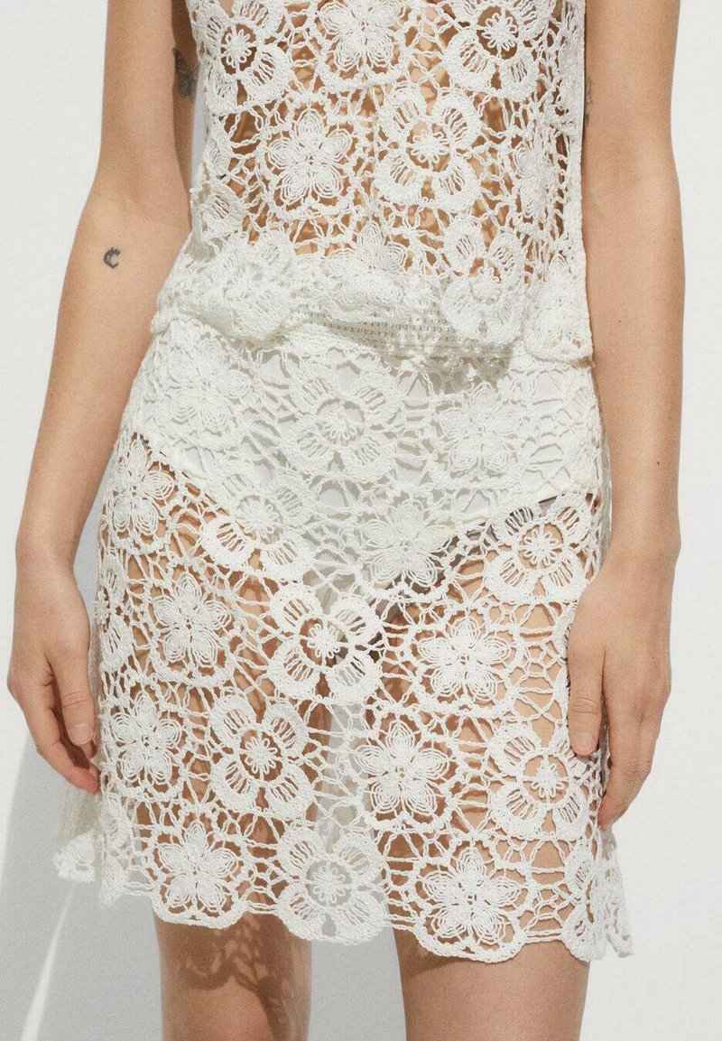 Rochie albă din croșetă cu modele florale, textură transparentă și tiv evazat. Dantela elaborată creează un design deschis pe întreaga suprafață.