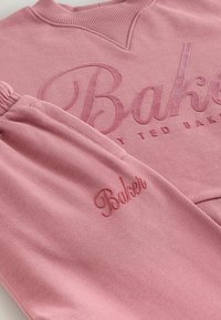 Rožinė džemperis ir atitinkamos sportinės kelnės su užrašyta "Baker by Ted Baker" toninėmis rožinėmis siūlėmis ant krūtinės ir šlaunų.