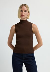 Vrouw met kort blond haar, grote oorbellen, een mouwloze bruine ribgebreide turtleneck en donkerblauwe jeans voor een effen achtergrond.