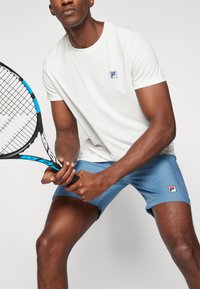 White short-sleeve athletic shirt with a blue logo, paired with sky-blue shorts. Holding a black tennis racket with blue accents.
