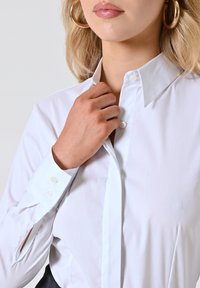 Camicia bianca a maniche lunghe con colletto a punta, polsini bottonati e texture liscia. Il modello regola il colletto con una mano.