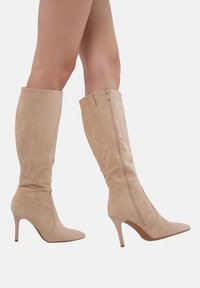Botas altas de ante color beige con punteras afiladas y tacón de aguja. Cuentan con un cierre lateral y una textura suave.