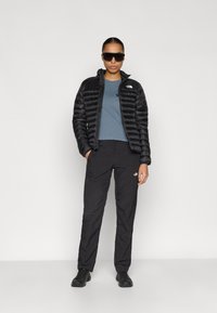 Μαύρο μπουφάν puffer με ψηλό γιακά, μπλε κοντομάνικο πουκάμισο και μαύρο παντελόνι, σε συνδυασμό με μαύρα γυαλιά ηλίου και μαύρα παπούτσια.