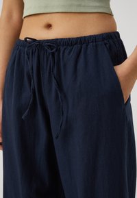 Närbild av en person som bär marinblå dragkedjebyxor med händerna i fickorna och en kort olivegrön topp.