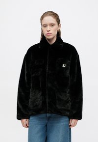 Chaqueta de pelaje sintético negro con cuello en punta, dos bolsillos frontales y un pequeño parche con logo en el pecho. Textura suave y corte relajado.