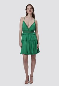 Vestido verde sin mangas con un profundo escote en V, detalles de encaje y dobladillo con volantes. Hecho de una tela ligera, presenta una cintura entallada y una falda acampanada.