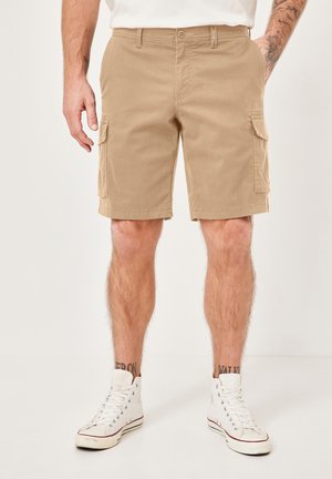 Man wearing beige cargo shorts with side pockets, white high-top sneakers, and a white t-shirt, standing with hands in pockets.