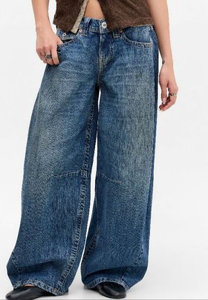 Osoba nosząca szerokie, wysokie na talii niebieskie jeansy z denimu z fakturowanym wzorem i czarne buty, trzymająca jedną rękę w kieszeni.