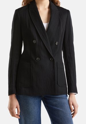Zwarte double-breasted blazer van een gemêleerde stof, met geknipte revers, twee voorzakken en zwarte knopen. Gedragen over een witte top en jeans.