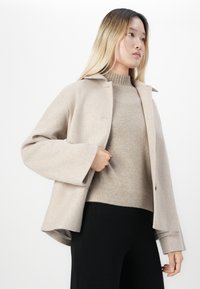 Woman wearing a beige wool coat over a beige knit sweater and black pants, standing against a white background.