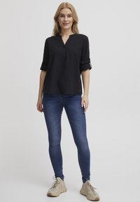 Tunic nera in lino con colletto alla cinese, maniche a tre quarti arrotolate, dettagli con bottoni, abbinata a jeans skinny blu e sneakers beige.