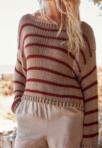 Femme portant un pull en maille beige avec des rayures horizontales rouges et un short beige clair, debout en plein air avec les mains dans les poches.