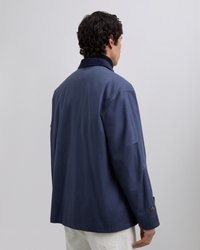 Mann mit dunklem lockigem Haar, der eine marineblaue Jacke und weiße Hosen trägt, mit dem Rücken zu einer schlichten hellen Hintergrund.