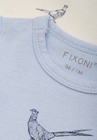 Ljusblå bomullsskjorta med rund nacke och korta ärmar. Har ett svart fågeltryck och grå sömmar längs kragen. Varumärkesetikett synlig.