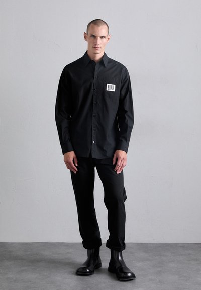 Cămașă neagră cu nasturi, cu un buzunar ce prezintă un cod de bare. Asortată cu pantaloni negri slim-fit și cizme până la glezna. Material din bumbac neted.