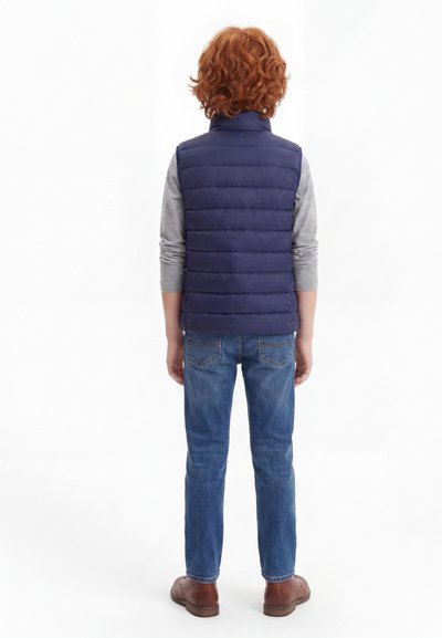 Παιδί με κόκκινα σγουρά μαλλιά που φοράει μια σκούρα μπλε επενδυμένη γιλέκο πάνω από γκρι μακρυμάνικη μπλούζα, μπλε τζίν και καφέ παπούτσια, στέκεται με την πλάτη γυρισμένη.