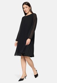 Czarna plisowana sukienka z długimi, przezroczystymi rękawami i okrągłym dekoltem. Do tego czarne mule na szpic o gładkiej fakturze.