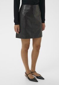 Crna kožna suknja A-linije, duljine do sredine bedra, glatke teksture, s minimalnim šavovima, uparena s crnim balerinkama s uskim remenom oko gležnja.