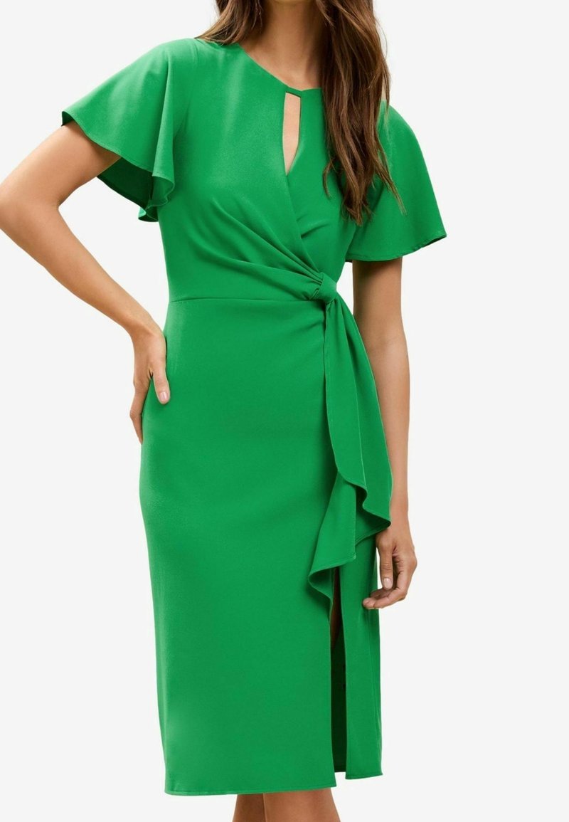 Femme portant une robe mi-longue vert vif avec des manches volantées, une fente frontale, un col en trou de serrure et un détail noué à la taille.