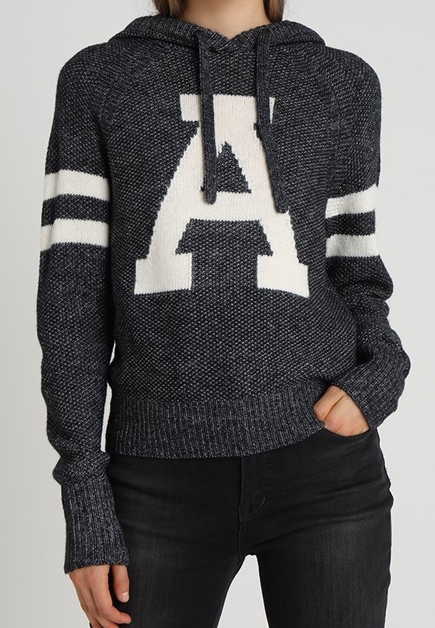Personne portant un sweat à capuche en tricot gris foncé avec des rayures blanches sur les manches et une grande lettre blanche "A" sur le devant, assorti à un pantalon noir.