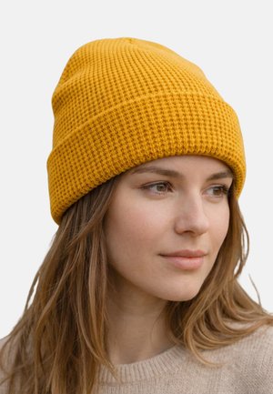 Junge Frau mit einer strukturierten, gelben Strickmütze, langen braunen Haaren und einem beigen Pullover, die leicht nach rechts schaut.