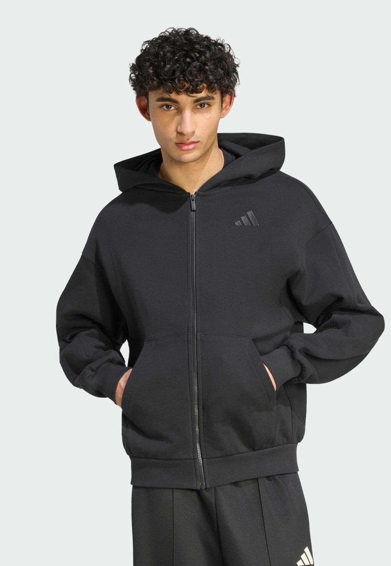 Jeune homme aux cheveux bouclés portant un sweat à capuche noir zippé et un pantalon noir assorti, les mains dans les poches du sweat.