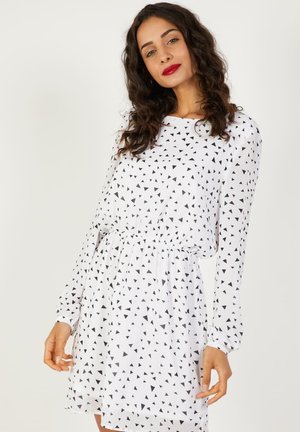 Mujer con cabello rizado y lápiz labial rojo, vestida con un vestido blanco de manga larga cubierto de pequeños patrones de triángulos negros, de pie frente a un fondo blanco.