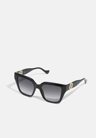 Zwarte zonnebril met vierkante montuur, grijze gradientlenzen en gouden Gucci-logo op de brillenpootjes, op een witte achtergrond.