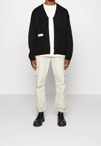 Cardigan de punto negro con apertura frontal, bolsillos laterales y puños acanalados, llevado sobre una camiseta blanca y pantalones cargo beige claro, combinado con botas negras.