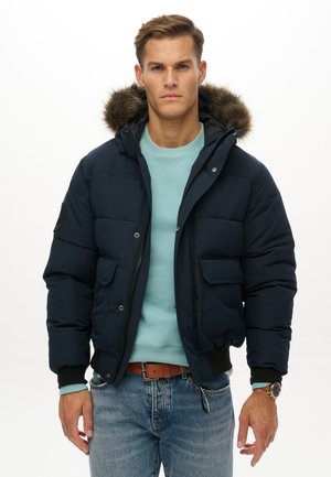 Mężczyzna w granatowej kurtce puchowej z kapturem wykończonym futerkiem, ubrany w jasnoniebieski sweter i dżinsy, stoi na białym tle.