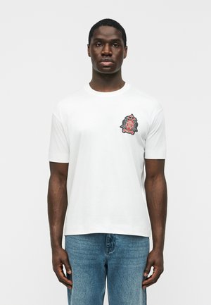 Férfi, aki fehér rövid ujjú pólót visel, amelynek bal mellén egy piros-fekete címerezett embléma látható, kék farmernadrággal, egyszínű háttér előtt.