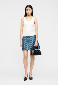 Γυναίκα με λευκή αμάνικη μπλούζα και μπλε φούστα με σχέδια, κρατώντας μικρό μαύρο τσαντάκι, στέκεται μπροστά σε λευκό φόντο.