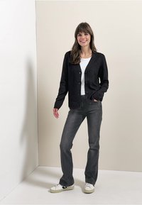 Černý cardigan, zapínání na knoflíky, texturovaná tkanina; v kombinaci s bílým tričkem a šedými džíny. Doplňují ho pohodlné sneakers. Neutrální pozadí.