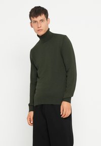 Pulover din tricot, de culoare verde închis, cu guler înalt; are mâneci lungi și o siluetă ajustată; asortat cu pantaloni negri.