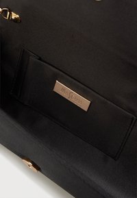 Wnętrze czarnej torebki pokazujące małą kieszonkę z złotą prostokątną plakietką marki z napisem "MASCARA LONDON" oraz złotym magnetycznym zapięciem.