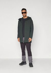 Tamsiai žalia neperšlampama striukė su gobtuvu, derinama su tamsiai pilkomis kelnėmis, turinčiomis šviesesnio pilko atspalvio akcentų. Juodi sportiniai batai ir saulės akiniai.