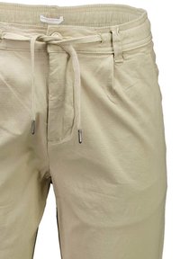 Pantalons en coton beige avec une taille à cordon de serrage, poches avant et accents métalliques sur les extrémités du cordon. Texture lisse, design décontracté.