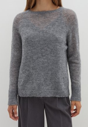 Vrouw die een zachte, pluizige grijze trui met lange mouwen en losse donkergrijze broek draagt, getoond van nek tot heupen tegen een effen achtergrond.