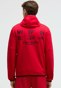 Sudadera roja con capucha, que presenta texto y gráficos negros en la parte posterior, y un acento negro en los puños y el dobladillo.