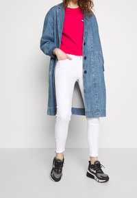 Abrigo de denim con grandes botones, mangas largas y bolsillos laterales sobre una camiseta rosa. Pantalones blancos ajustados con cremalleras laterales y zapatillas deportivas negras.